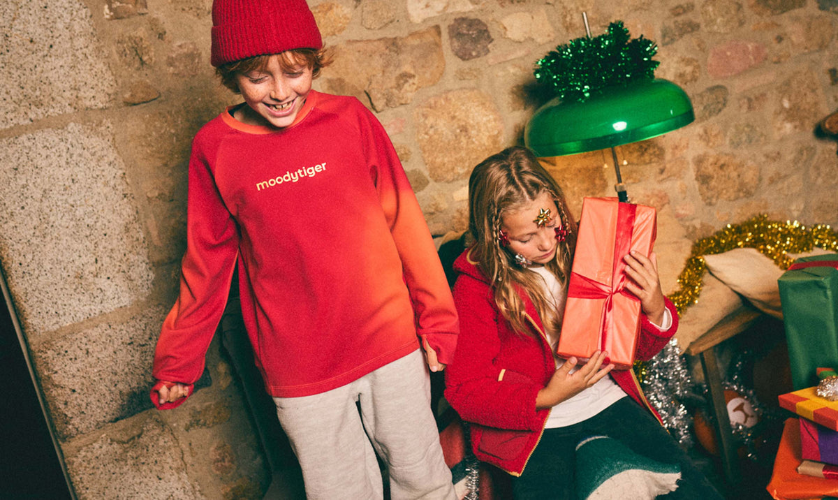 Two kids in red moodytiger tops holding wrapped gifts by a festive stone wall