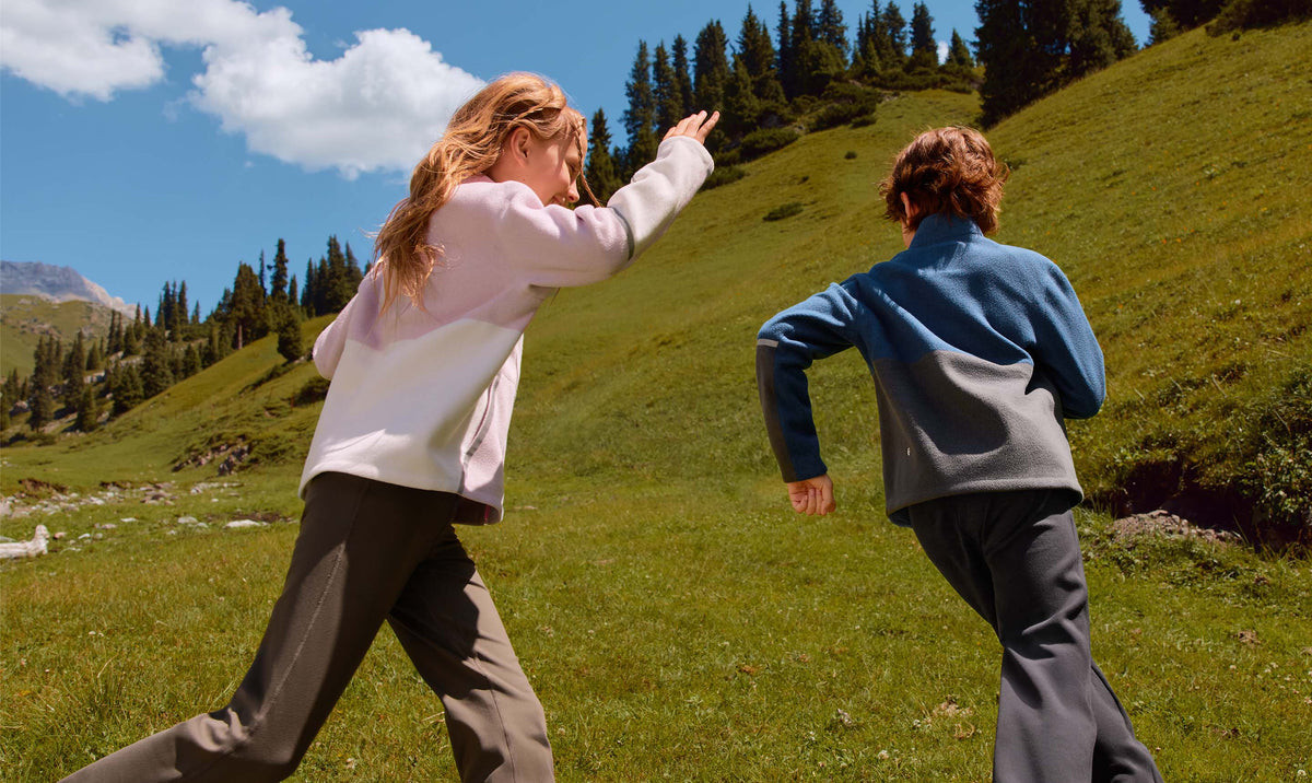 Girl in a pink fleece jacket and boy in a blue colour-block fleece jacket running up a green alpine meadow